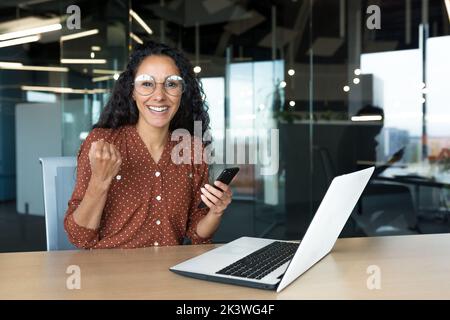 Portrait d'une femme d'affaires latino-américaine réussie, d'une travailleuse en lunettes et de cheveux bouclés regardant l'appareil photo et souriant heureux célébrant le triomphe et gagnant assis à une table tenant le téléphone. Banque D'Images