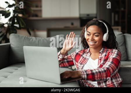 Femme afro-américaine souriante du millénaire dans un casque sans fil étudiant avec un ordinateur portable, en agitant la main Banque D'Images
