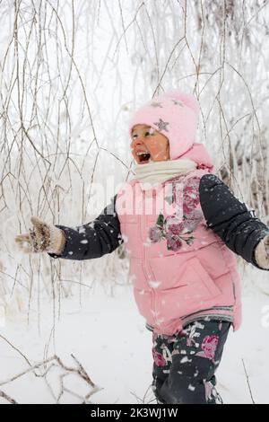 Petite fille aux joues rudes et large sourire extrêmement heureux dans les vêtements chauds d'hiver courir sur la neige avec des arbres neigeux en arrière-plan. Promenade en hiver Banque D'Images