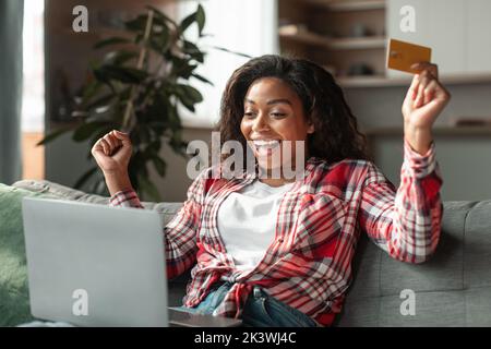 Satisfaite surprise millénaire noire dame avec ordinateur portable et carte de crédit se réjouit dans la victoire et le succès des achats en ligne Banque D'Images