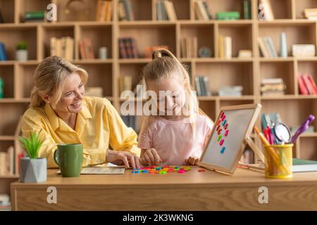 Joyeuse petite fille d'école maternelle étudiant à lire avec un tuteur professionnel, positive femme aidant l'enfant à apprendre des lettres Banque D'Images