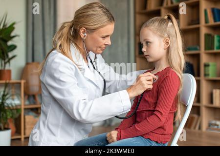 Femme médecin vérifiant les poumons de l'enfant pendant le contrôle médical à l'intérieur de la maison, en utilisant le stéthoscope pour examiner la respiration Banque D'Images