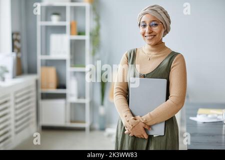 Portrait minimal de la jeune femme d'affaires du Moyen-Orient souriant à l'appareil photo dans un bureau et tenant un ordinateur portable, un espace de copie Banque D'Images