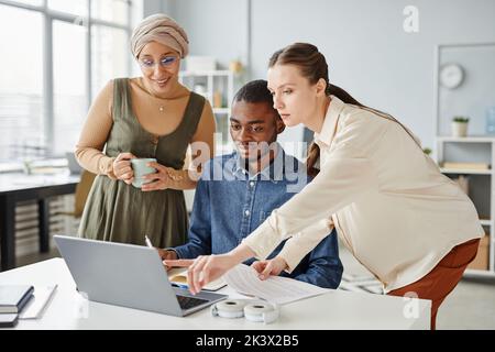 Groupe diversifié de trois professionnels utilisant un ordinateur portable ensemble tout en discutant du projet dans un bureau minimal Banque D'Images