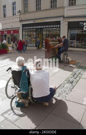 Ville de Bath, Somerset Angleterre UK.Male Street Busker jouant du piano dans la High Street. Femme en fauteuil roulant accompagnée d'un compagnon masculin regardant sur. Banque D'Images