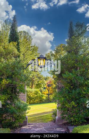 Belle porte de jardin formelle avec lanterne et beaucoup de feuillage menant à la prairie ensoleillée avec des arbres à distance Banque D'Images