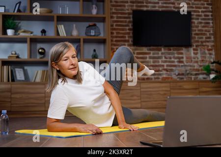 Femme mûre heureuse à cheveux gris faisant de la gymnastique et du yoga à la maison en ligne avec un ordinateur portable. Un mode de vie actif et sain pour les retraités. Cours de fitness Banque D'Images