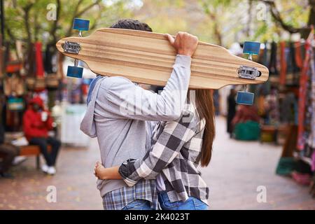 Prendre un temps de l'embarquement. Photo amusante d'un jeune couple qui embrasse derrière un skateboard. Banque D'Images