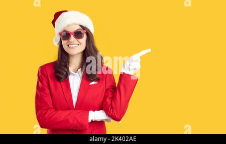 Belle femme en chapeau de père noël et costume rouge pointant avec le doigt sur l'espace de copie sur fond orange. Banque D'Images