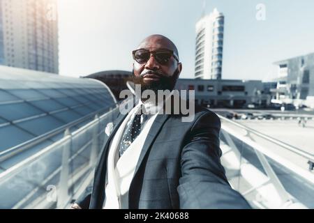 Un homme noir à tête blanche et barbu prend un selfie de première personne dans la rue ; un homme d'affaires africain à la mode en costume taillé sur mesure prend le pas Banque D'Images