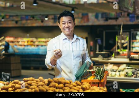 Un jeune homme d'affaires asiatique beau est debout dans un supermarché de la section des légumes, tenant un panier de fruits et de pommes de terre dans ses mains. Il regarde la caméra, sourit. Banque D'Images