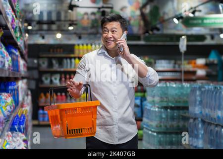 Beau jeune homme asiatique marche dans le supermarché entre les étagères avec un panier orange pour les produits, parle au téléphone, choisit, sourit. Banque D'Images