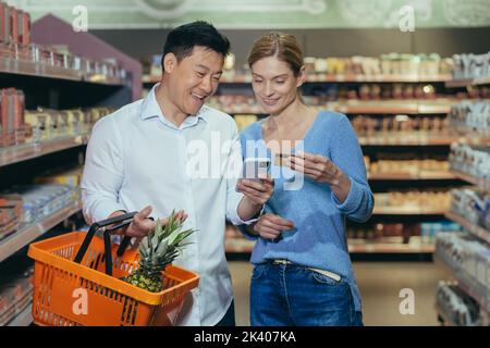Shopping en famille. Jeune beau couple interracial shopping dans un supermarché. Femme et homme asiatiques heureux tenant la carte de crédit et le téléphone, payant sans espèces, faisant la commande en ligne. Banque D'Images