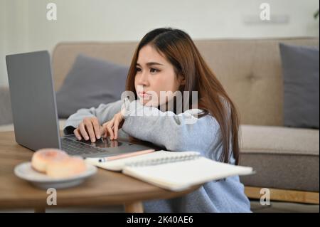 Une jeune femme asiatique ennuyeuse, somnolente et fatiguée fait ses devoirs dans le salon, recherchant des informations en ligne sur sa comput portable Banque D'Images