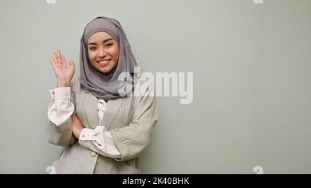 Jeune femme musulmane asiatique souriante et belle, portant le hijab, se tenant isolée sur fond de studio gris. Salutation à la main par vague Banque D'Images