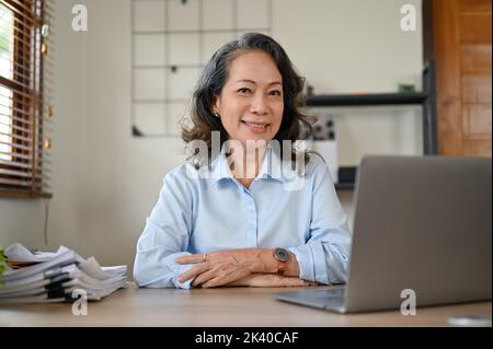 Femme d'affaires ou directrice senior asiatique d'âge prospère et professionnelle assise à son bureau, souriant et regardant la caméra. Banque D'Images