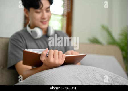 Un jeune homme asiatique élégant et charmant lisant un livre ou un manuel tout en se relaxant sur un canapé dans son salon. Attention sélective Banque D'Images