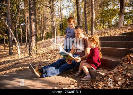 papa est en train de lire un livre pour les enfants dans le parc. en lisant un livre pour un fils et une fille dans le jardin d'automne Banque D'Images