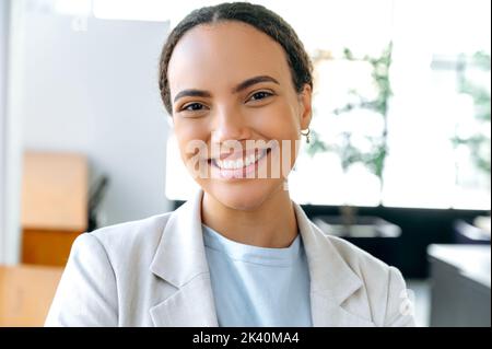 Photo en gros plan d'une femme brésilienne ou hispanique confiante et charmante, en tenue formelle, femme d'affaires, directrice de la société, se tient au bureau, regarde la caméra, avec un sourire sincère et positif Banque D'Images
