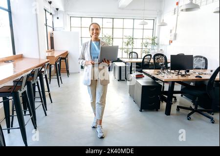 Jeune femme de course mixte moderne et confiante, vêtue de vêtements de travail élégants, traverse le bureau créatif en tenant un ordinateur portable ouvert entre ses mains, en regardant l'appareil photo, en souriant Banque D'Images