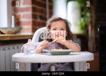 portrait petite fille caucasienne d'environ 2 ans dans un bavoir mangeant des pâtes à partir d'une assiette assis chaise haute, auto-alimentation pour les enfants, idée de sevrage menée par bébé Banque D'Images