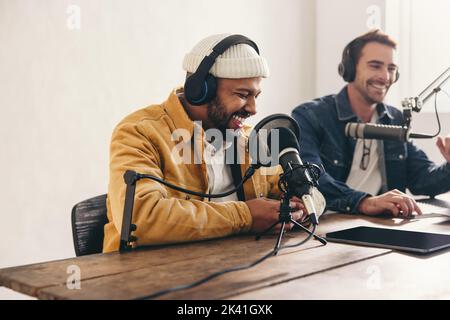 Deux podcasters d'université rient et ont un bon moment dans un studio. Deux jeunes hommes heureux co-accueillent une émission audio en direct. Deux créateurs de contenu de sexe masculin r Banque D'Images