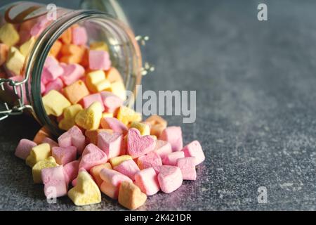 Tas de bonbons guimauves en forme de coeur rose et jaune, dispersés dans un pot en verre sur fond gris avec espace de copie Banque D'Images