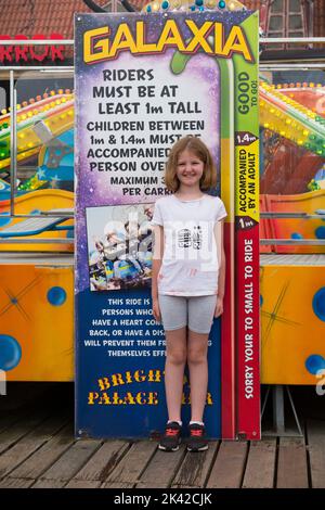 Fille enfant 1,4 m mètres grande fille enfant / huit fille de 8 ans debout à une échelle de mesure de la hauteur qui indique qu'elle est assez grande pour aller sur une hauteur limitée fairground tour / foire avec des divertissements, attractions, Et des promenades incluant la Galaxia avec sa taille limitée, au bout de Brighton Palace Pier. ROYAUME-UNI. (131) Banque D'Images