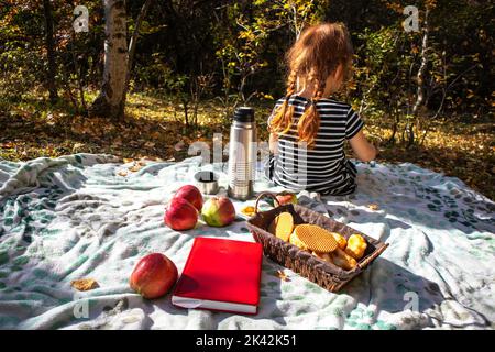 une petite fille aux cheveux rouges dans une robe rayée est assise avec son dos sur un pique-nique en automne. Banque D'Images