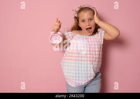 Concept enfants. Une fille tient un réveil dans ses mains, joue avec elle, regarde la caméra. Isolé sur fond rose. Banque D'Images