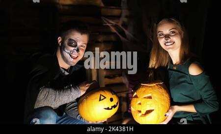 Date dans le style de la fête d'Halloween, nuit, crépuscule, dans les rayons de lumière, un gars avec une fille vêtue en costumes et avec un maquillage terrible tiennent deux grandes citrouilles d'Halloween. Photo de haute qualité Banque D'Images