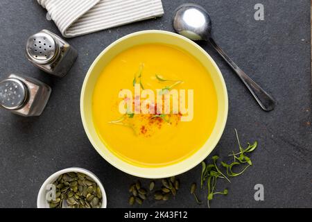 Image horizontale d'un bol de soupe de carottes avec garniture, condiments, cuillère et graines de citrouille sur ardoise Banque D'Images