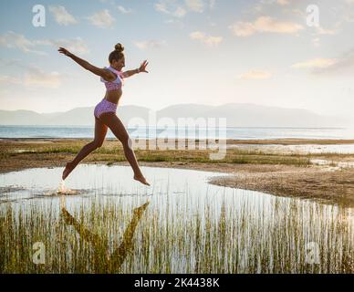 Adolescente (14-15) au bikini sautant au-dessus du lac au lever du soleil Banque D'Images