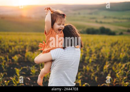 Mère gaie avec sa petite fille insouciante dans ses bras, s'amuser et jouer tout en marchant dans le champ de maïs au coucher du soleil. Le mode de vie des gens Banque D'Images