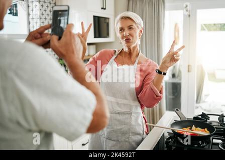 Un couple senior qui fait la cuisine, un smartphone qui prend le portrait en cuisine et une femme avec le signe de paix v pour les médias sociaux, la célébration de la retraite ou la santé Banque D'Images