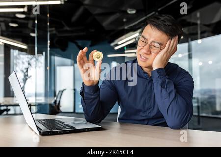 Frustré investisseur d'homme d'affaires asiatique tenant la crypto-monnaie bitcoin dans les mains, homme travaillant dans un immeuble de bureaux moderne, échec avec l'investissement financier. Banque D'Images