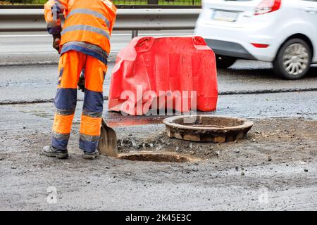 Un ouvrier de la route nettoie la bouche d'un trou d'égout sur la chaussée contre le fond d'une voiture qui passe dans le flou. Banque D'Images