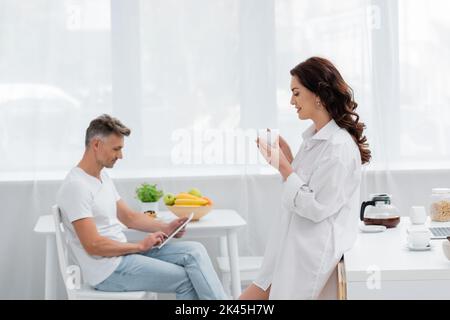 Vue latérale d'une femme souriante dans une chemise tenant un café près d'un mari flou utilisant une tablette numérique dans la cuisine, image de stock Banque D'Images