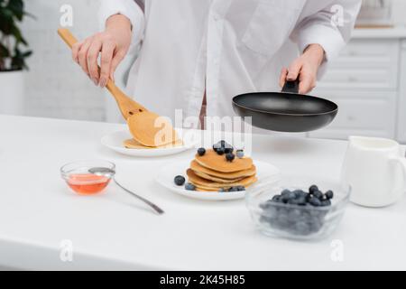 Vue rognée de la femme en chemise mettant la crêpe sur l'assiette près des bleuets flous dans la cuisine, image de stock Banque D'Images