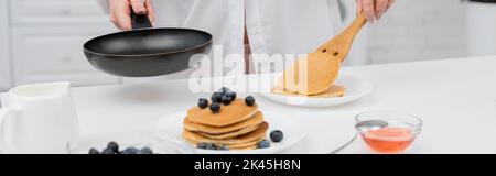 Vue rognée de la femme en chemise mettant la crêpe sur l'assiette près des bleuets frais dans la cuisine, bannière, image de stock Banque D'Images