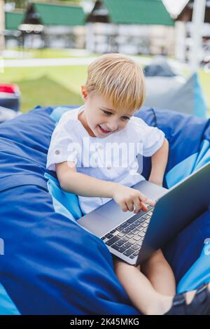 Petit garçon avec un ordinateur portable sur le fond de l'herbe d'été. Un garçon dans un T-shirt blanc est assis sur un sac de beanbag dans le parc. Banque D'Images