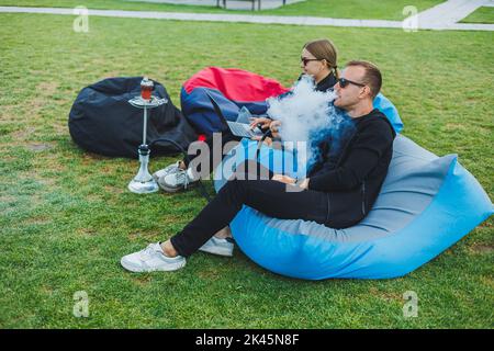 Un jeune couple se détend dans le parc et fume le narguilé. Un homme et une femme s'assoient sur des chaises dans la nature et fument un narguilé. Vacances en famille Banque D'Images