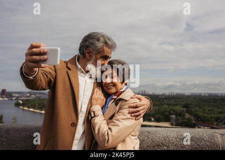 homme senior heureux embrassant la tête de femme gaie dans un manteau de tranchée et prenant selfie, image de stock Banque D'Images