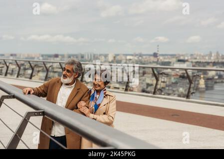 homme senior heureux en manteau et femme gaie debout près du rail de garde de pont, image de stock Banque D'Images