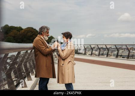vue latérale d'une femme âgée heureuse tenant les mains avec un mari barbu et gai près du rail de garde-pont, image de stock Banque D'Images