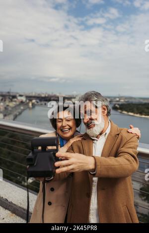 homme senior heureux prenant le selfie avec une femme gaie sur un appareil photo vintage avec ville floue sur l'arrière-plan, image de stock Banque D'Images