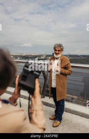 femme floue tenant un appareil photo d'époque tout en prenant la photo d'un mari aîné barbu sur le pont près de la rivière, image de stock Banque D'Images