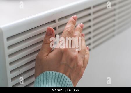 Gros plan d'une femme en chandail réchauffe ses mains sur la batterie sur fond de mur blanc. Photo macro des mains de la femme, espace de copie. Concept de Banque D'Images