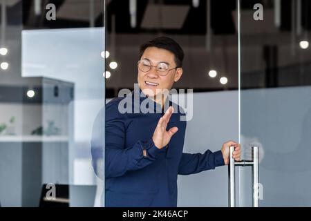 L'homme entre dans la salle de réunion, sourit et salue, le patron asiatique en lunettes et chemise, l'investisseur d'homme d'affaires travaille dans un immeuble de bureaux moderne Banque D'Images
