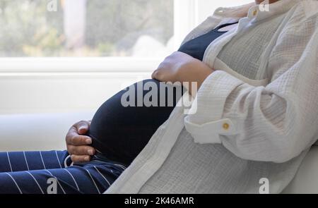 Vue rapprochée d'une femme enceinte assise sur un canapé près de la fenêtre - grossesse, attente, concept de maternité Banque D'Images